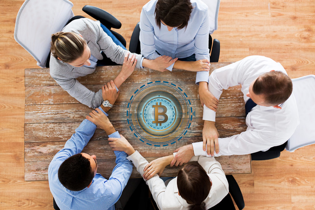 cryptocurrency, cooperation and finance concept concept - business team sitting at office table with bitcoin hologram and holding hands from top